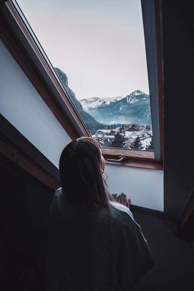 Eine Frau steht innen vor einem Dachfenster und schaut auf eine verschneite Landschaft