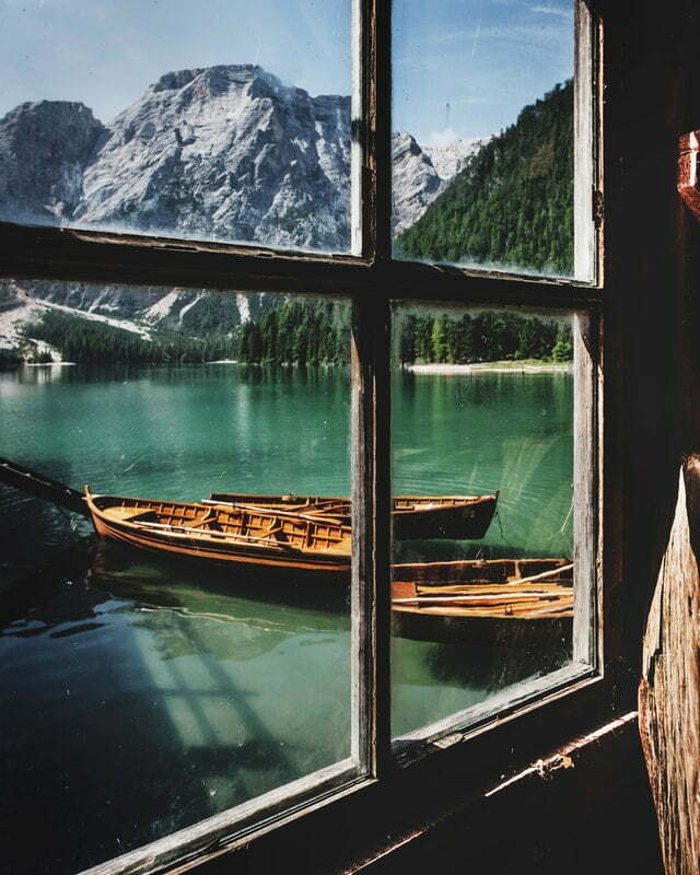 Aus einem einfachverglastem Fenster schaut man auf einen See mit Bergen im Hintergrund
