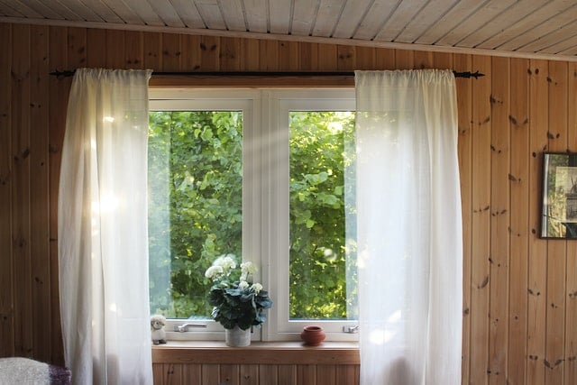 Vor einem Fenster, welches in eine Holztäfelung eingelassen ist, hängt en weißer Vorhang