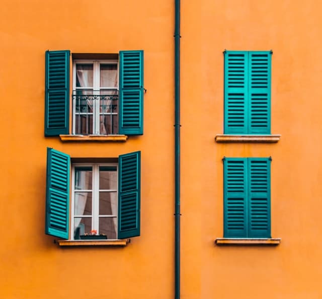 An einer orangenen Wand liegen Fenster mit türkisen Fensterläden