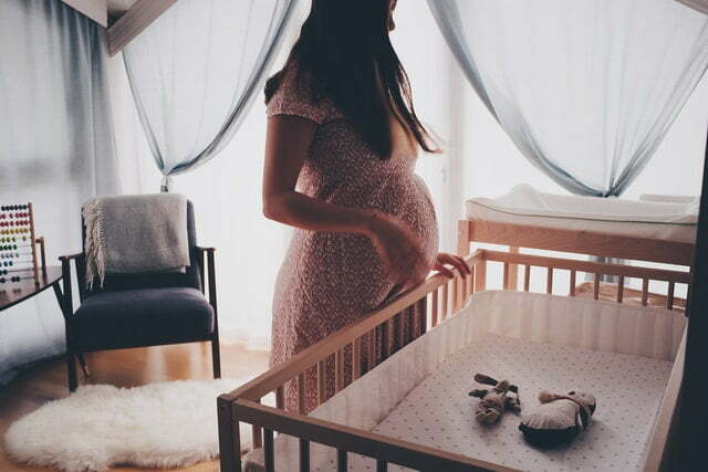 Eine schwangere Frau steht vor einer Krippe, dahinter gauzeartige Vorhänge