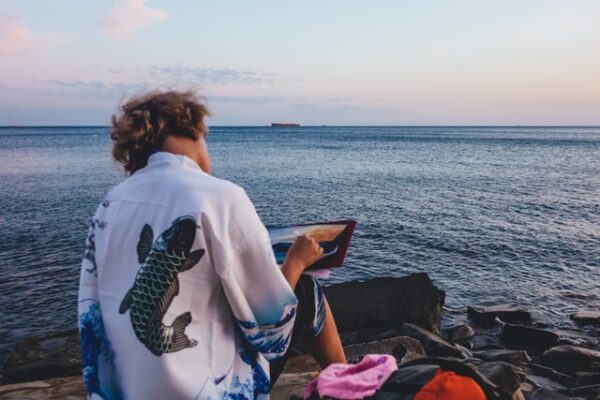 Ein Mann sitzt am Meer und zeichnet auf einem Tablet