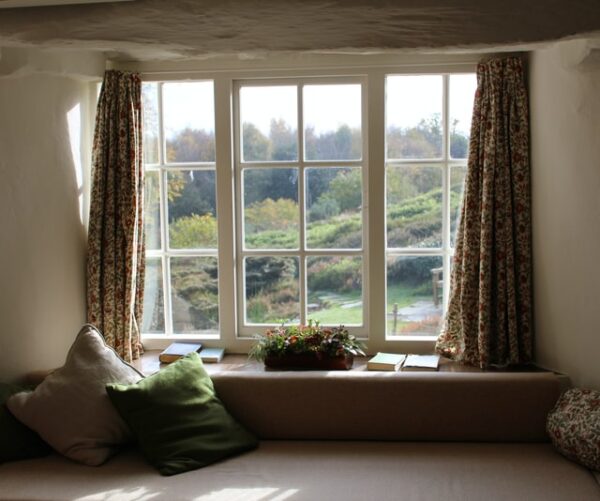 Ein Fenster zeigt ins Grüne. Davor befinden sich Vorhänge, eine Couch und eine Pflanze