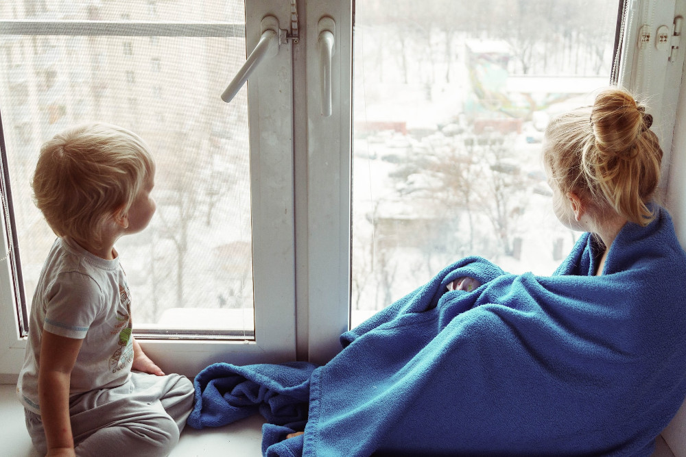 Zwei Kinder sitzen vor einem Fenster und schauen auf die verschneite Landschaft