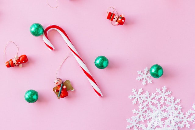 Auf einem rosa Untergrund liegen kleine weihnachtliche Ornamente in Form von Zuckerstangen, Schneeflocken und Kugeln