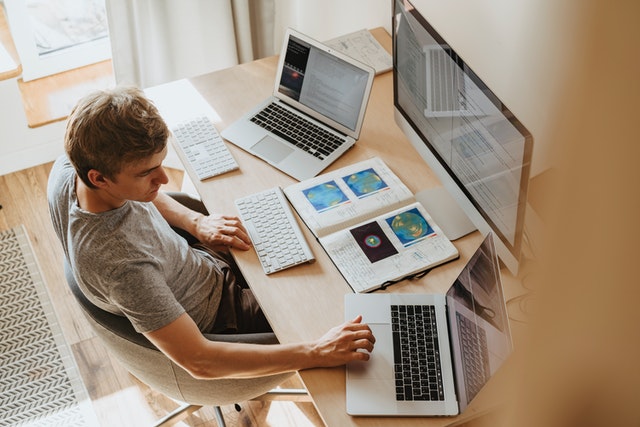 Ein Mann sitzt an einem Schreibtisch mit einem großen Bildschirm, zwei Notebooks und Notizbüchern