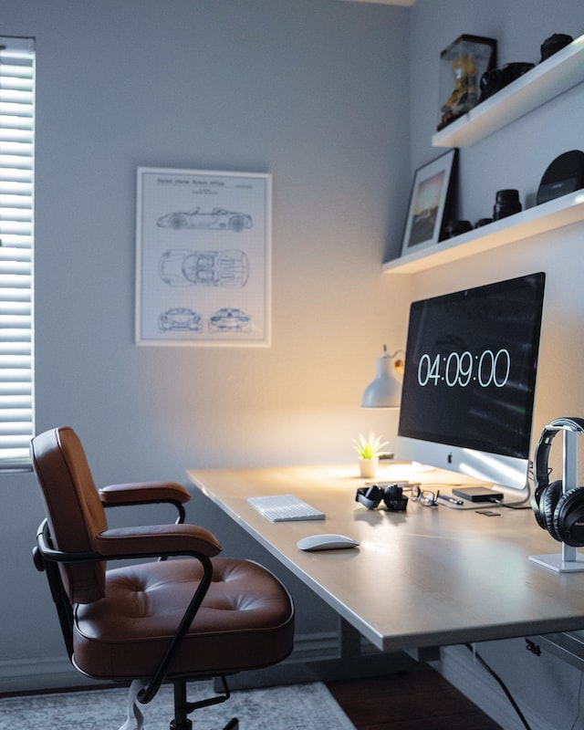Ein Schreibtisch in einer Ecke mit Stuhl, großem Bildschirm, Regalen und Bildern an der Wand