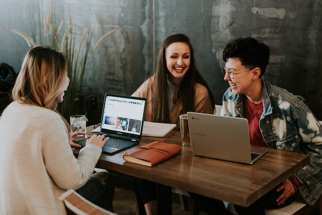 Drei Personen sitzen mit Laptops an einem Tisch und arbeiten lachend zusammen