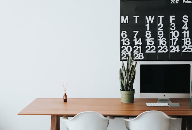 Auf einem Schreibtisch mit zwei Stühlen stehen Tastatur, Maus, eine Pflanze und an der Wand hängt ein Kalender