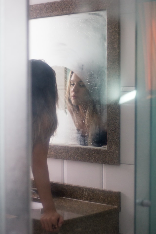 Eine Frau schaut ihre Reflexion in einem beschlagenen Spiegel an, auf dem sie vorher eine Stelle freigewischt hat