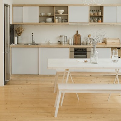 Heller Holzfußboden, darauf eine weiße Küche mit heller Holz-Arbeitsplatte, davor weite Tisch-Bank-Kombi