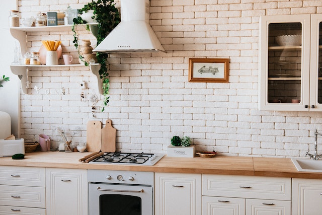 eine geweißte Ziegelsteinmauer vor der eine weiße Küche mit heller Holz-Arbeitsplatte steht
